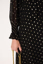 A person wearing a robe mousseline noire, sheer and long-sleeved with gold polka dots (pois lurex doré), holds a gold chain in their hand against a neutral background.