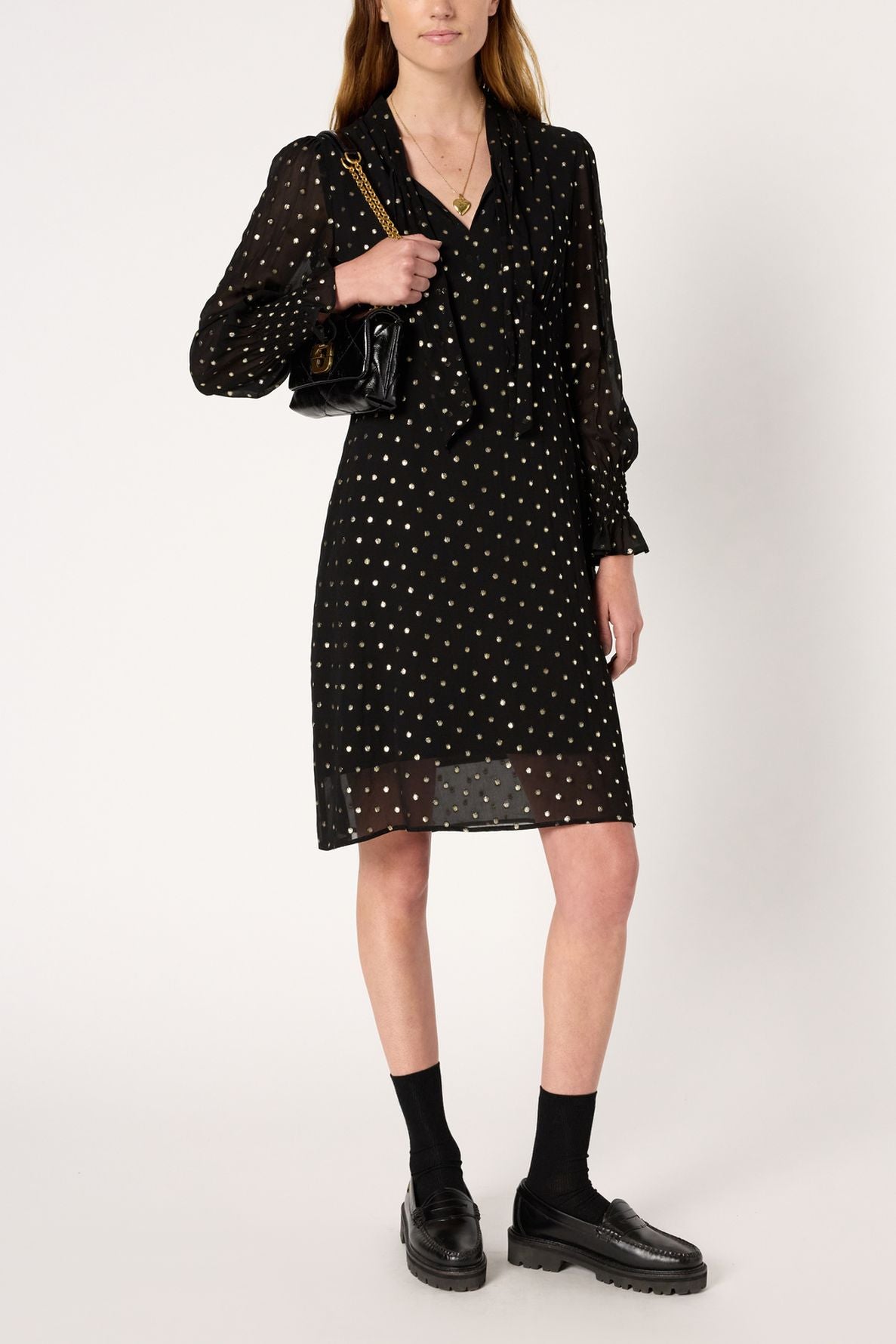 Woman wearing a black, long-sleeve, knee-length robe mousseline noire adorned with delicate pois lurex doré and featuring a chic col lavallière, paired with black socks and black loafers. She holds a black handbag with a chain strap while standing against a plain white background.