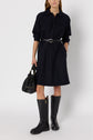 Woman wearing a black belted robe courte with col chemise and long sleeves, knee-high black boots, and holding a black handbag, standing against a plain light background.
