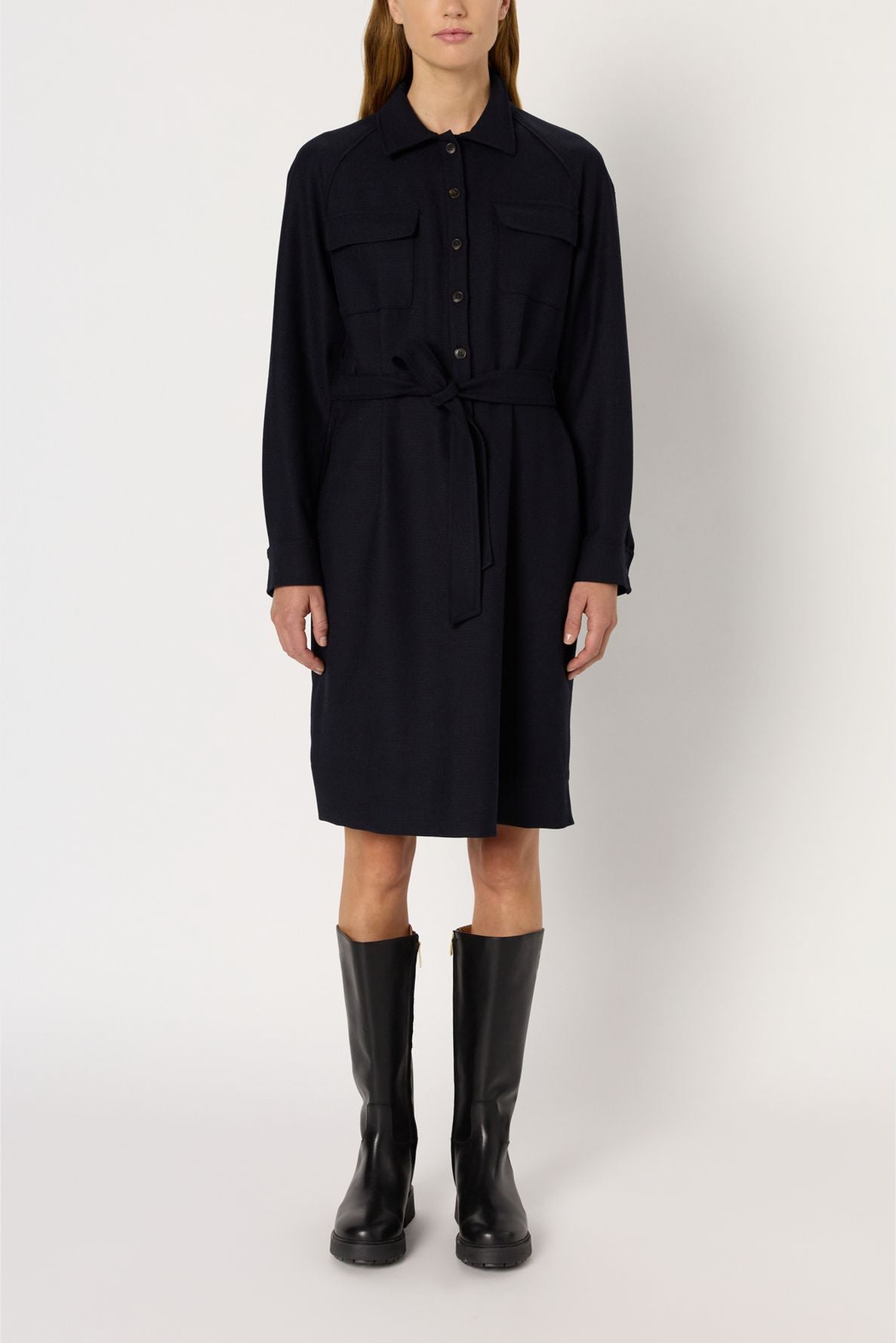 Person wearing a black, long-sleeve robe Léa style button-up shirt dress with a col chemise and two chest pockets, belted at the waist and paired with black knee-high boots, standing against a plain background.