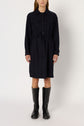 Person wearing a black, long-sleeve robe Léa style button-up shirt dress with a col chemise and two chest pockets, belted at the waist and paired with black knee-high boots, standing against a plain background.