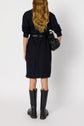 A person with long wavy hair, wearing a robe Léa and tall black boots, stands facing away. They carry a black bag over one shoulder.