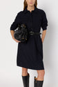 Woman wearing a robe courte style dark button-up dress with a col chemise and belt, holding a black handbag, and black knee-high boots. She is standing against a plain white background.