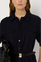 Woman wearing a dark col chemise button-up collared dress with chest pockets, a black leather belt with a gold buckle, and holding a black handbag with gold hardware. Her hair is down, and the background is plain.