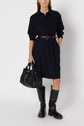A woman wearing a belted, long-sleeve, buttoned black robe Léa dress, black knee-high boots, and holding a large black handbag stands against a plain white background.