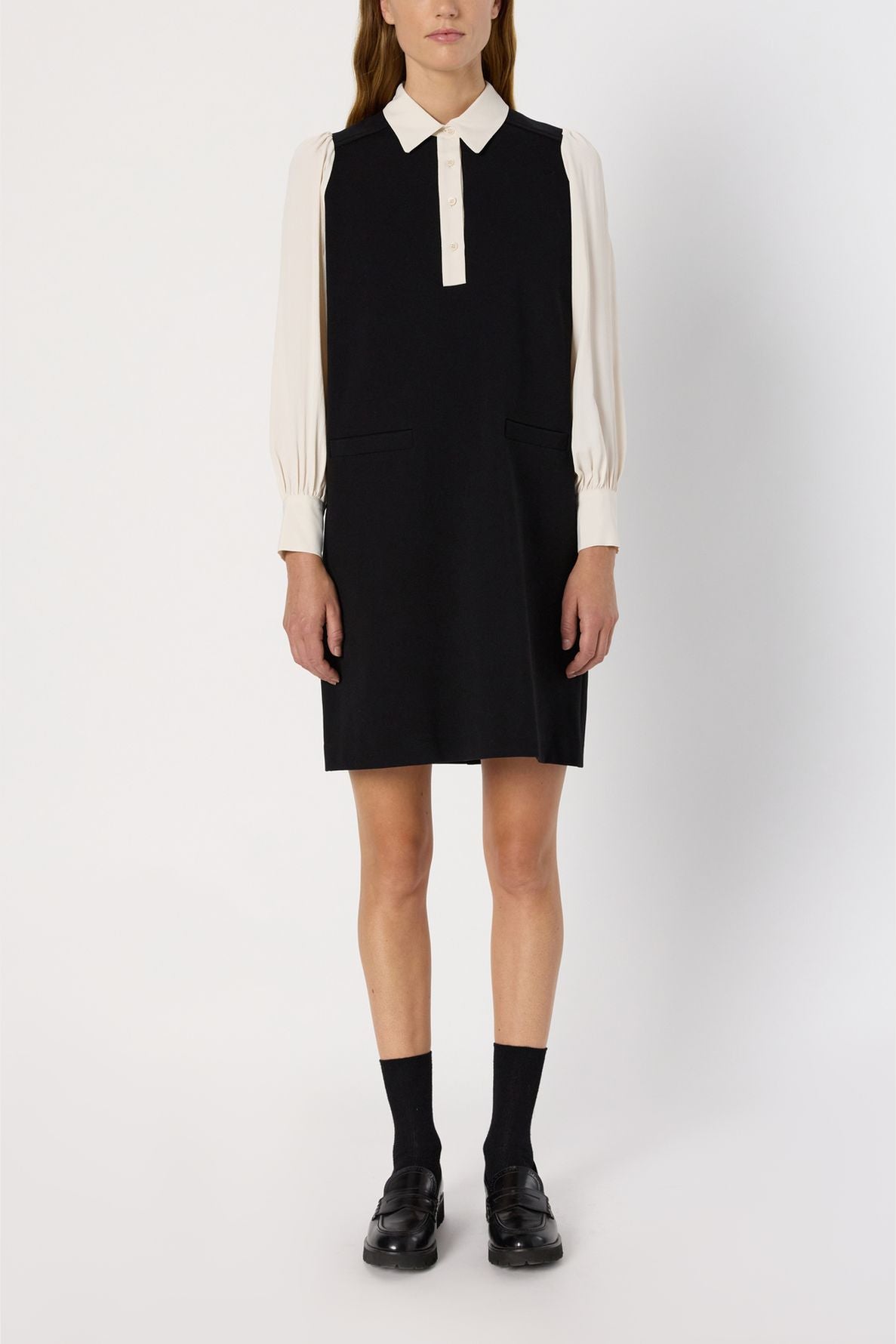 A person stands wearing a robe courte noire sans manches superposée sur un chemisier crème à col chemise, assortie de chaussettes noires et de chaussures noires, devant un fond blanc uni.