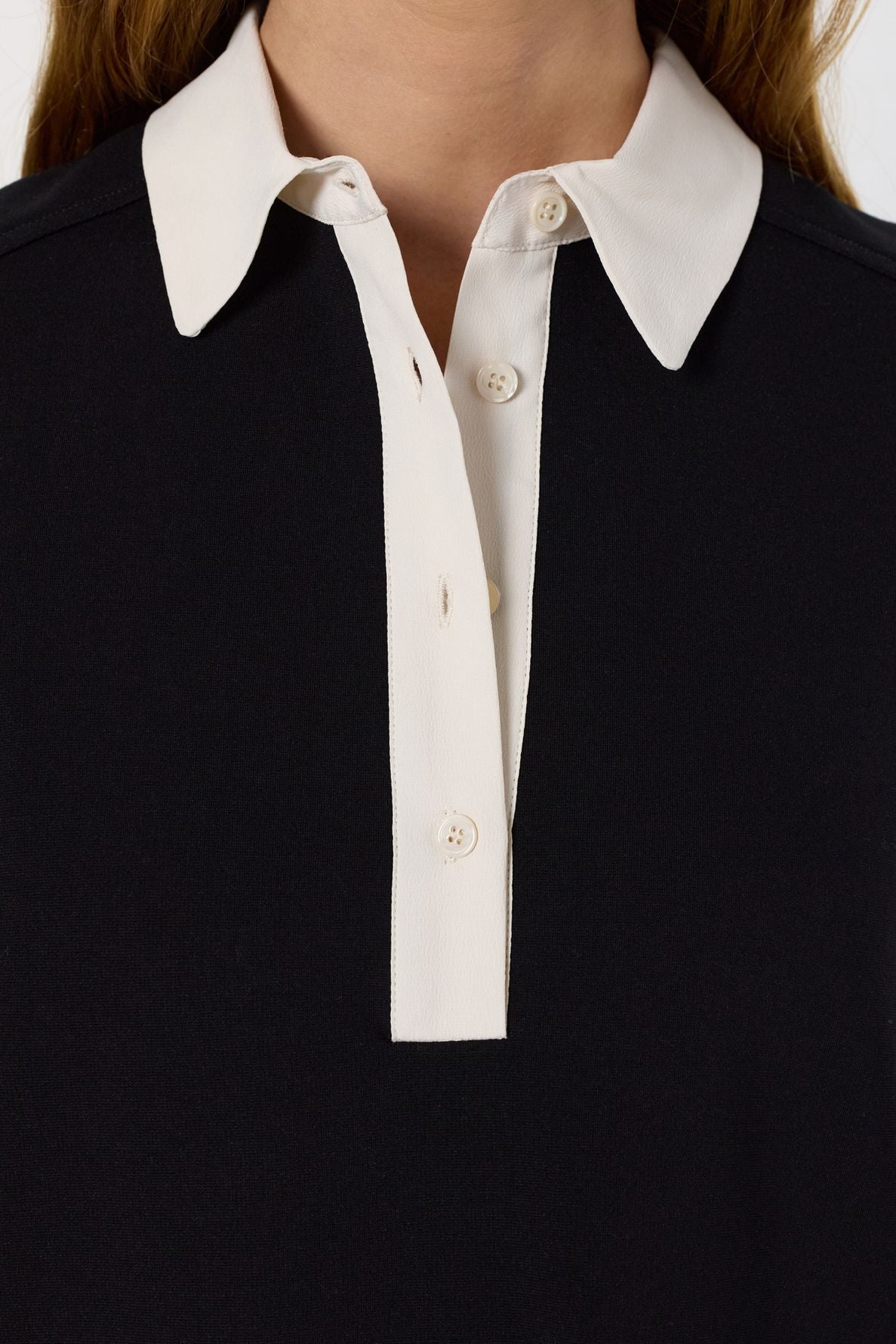 Close-up of a person wearing a black shirt with a white collar and white button placket, reminiscent of a col chemise style. The shirt has three visible buttons, and the wearer’s neck and part of the hair are visible.