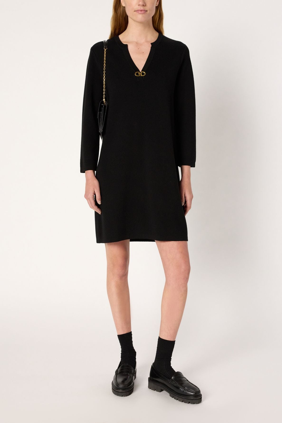 A person wearing a robe noire maille Milano, featuring a black long-sleeve design with a gold V-neck detail, paired with black loafers, black socks, and a small black shoulder bag, standing against a plain white background.