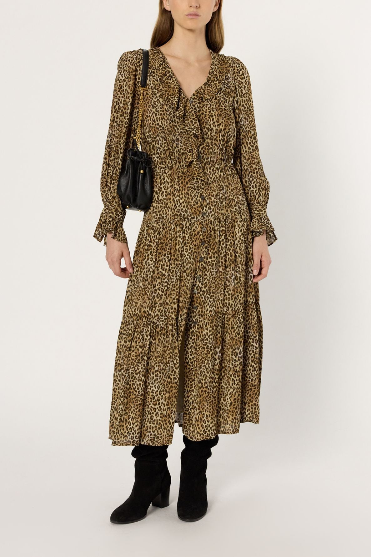 Woman in a long-sleeve, v-neck, ankle-length robe léopard with ruffles, black shoulder bag, and black knee-high boots standing against a plain white background.