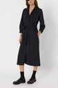 Woman wearing a dark, long-sleeved robe chemise midi with a ceinture à nouer at the waist, paired with black socks and black loafers. She stands against a plain white background.