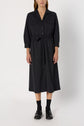 A woman wears a long-sleeve, button-front, charcoal grey robe chemise midi dress with two chest pockets and a ceinture à nouer at the waist. She pairs it with black socks and black loafers, standing against a plain white background.