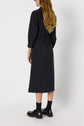A person with long, wavy blond hair wears a dark, long-sleeved, ankle-length robe chemise and black loafers with black socks, standing facing away from the camera against a plain white background.