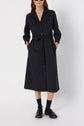 Woman wearing a dark jersey épais robe chemise midi with long sleeves, front pockets, a ceinture à nouer at the waist, black socks, and black loafers. She has her hands in the dress pockets and is standing against a white background.