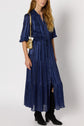 Woman wearing a robe longue, navy blue and pleated, with boutons, manches longues, and a cinched waist. She holds a gold purse and wears black knee-high boots, standing against a plain, light background.