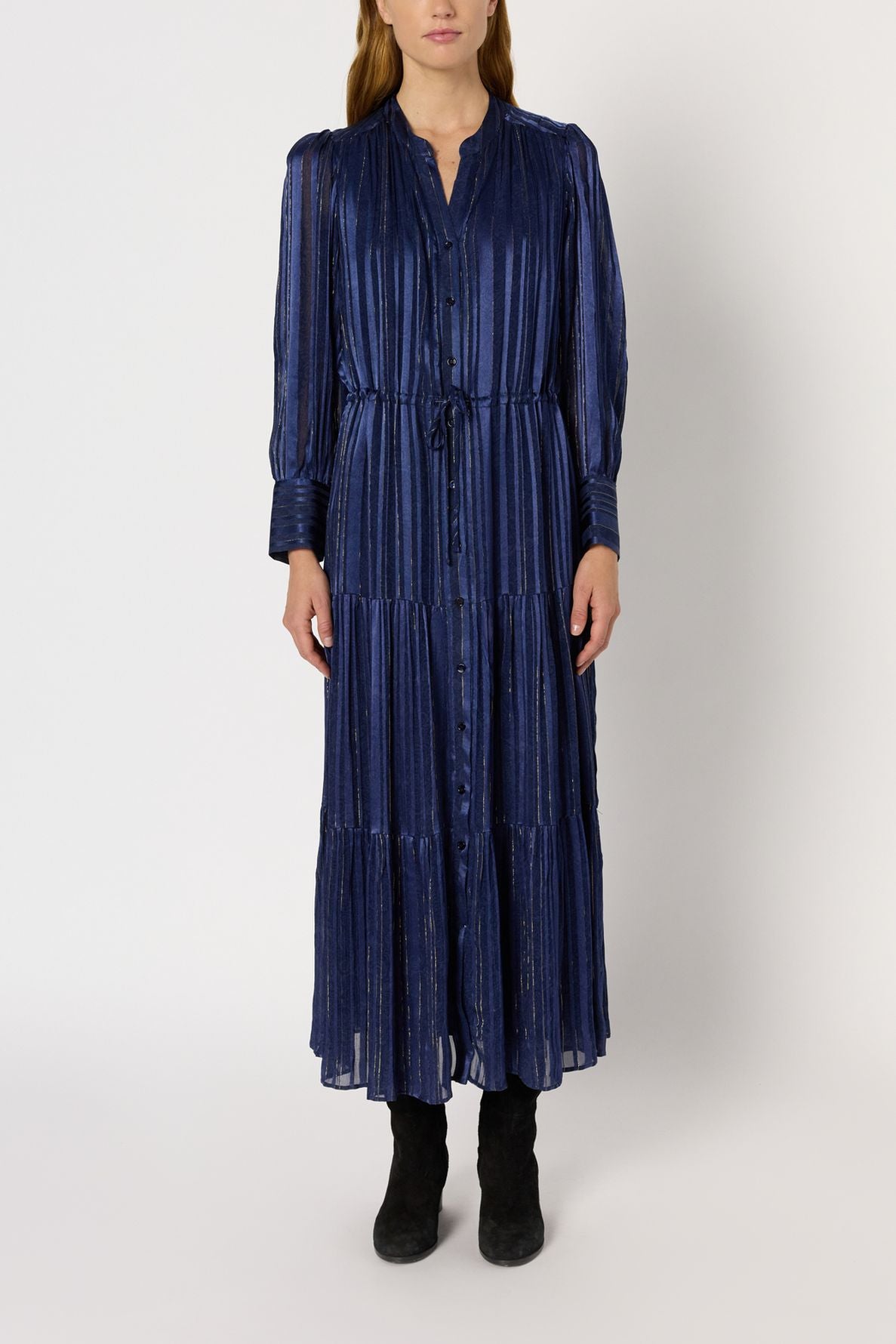 A person wearing a robe longue, manches longues, ankle-length, navy blue dress with vertical stripes and button details accented by subtle lurex doré threads, standing against a plain background and wearing black boots.