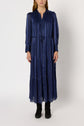 A person wearing a robe longue, manches longues, ankle-length, navy blue dress with vertical stripes and button details accented by subtle lurex doré threads, standing against a plain background and wearing black boots.