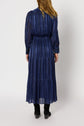 Woman with long blonde hair wearing a manches longues, ankle-length, dark blue striped robe longue with subtle lurex doré threads and black boots, standing with her back to the camera against a plain white background.