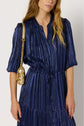 A woman in a blue, vertically striped robe longue with gold accents and manches longues holds a small lurex doré purse with a chain strap over her shoulder.