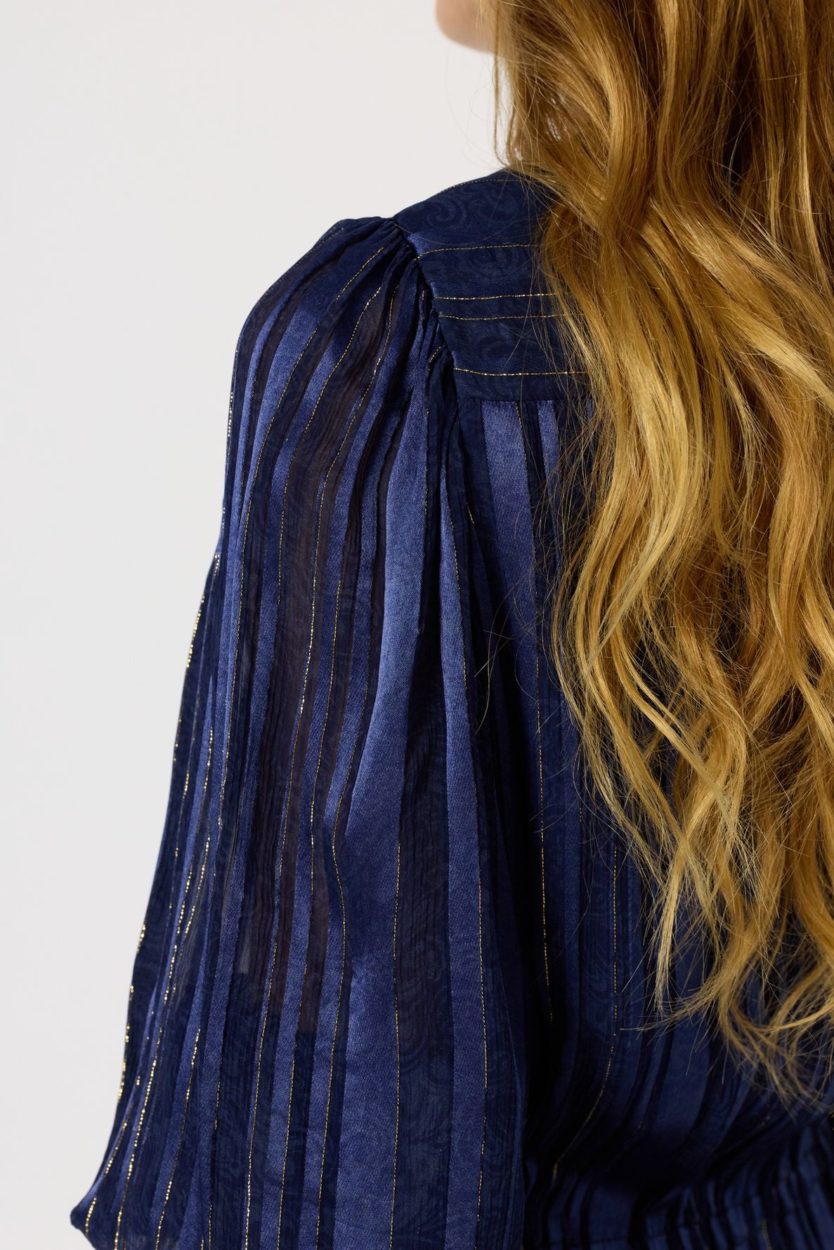 A woman with wavy blonde hair wears a navy blue, sheer, long-sleeve blouse with vertical stripes and subtle metallic lurex doré threads.
