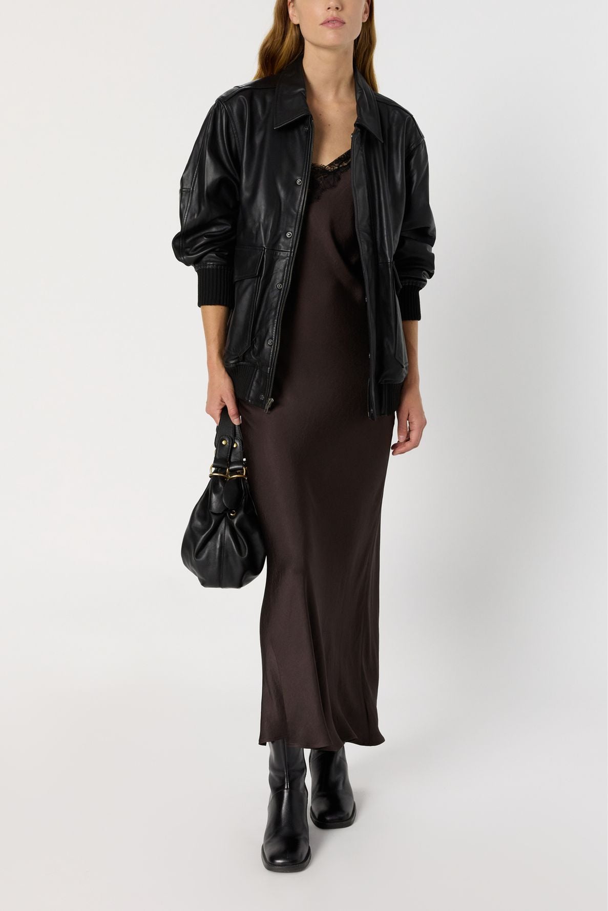 A woman in a black leather jacket, brown slip dress reminiscent of a robe longue satin, and black boots holds a black handbag. She stands against a plain white background, capturing the modern elegance of the collection Studio.