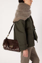 Person in a green jacket and brown pants with a thick brown scarf, carrying a dark brown besace cuir—a chic sac bandoulière with gold hardware—against a plain background.