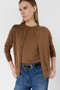 Woman wearing a brown cardigan over a brown top, blue high-waisted jeans with a black belt, standing with hands in pockets, looking to the side against a plain background.