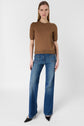 A woman stands against a plain background, wearing a brown short-sleeve knit top, blue wide-leg jeans, and black pointed-toe shoes. She faces forward with her arms relaxed by her sides.