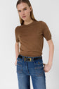 A woman with straight blonde hair wears a brown short-sleeve sweater, blue jeans with a black belt, and gold jewelry, standing with hands in her pockets against a plain background.