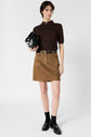 A woman stands against a white background, wearing a brown col rond short-sleeve turtleneck, tan mini skirt with pockets, black socks, black loafers, and holding a black handbag.