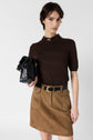 A woman in a brown laine mérinos top and tan skirt stands holding a black handbag. She has short dark hair and a neutral expression, with one hand in her pocket. The background is plain and light-colored.