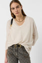 A woman with straight light brown hair wears a cream V-neck sweater, patterned gray jeans, a black belt with a gold buckle, a gold chain necklace, and a black shoulder bag, standing against a plain white background.