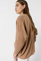 A woman with long brown hair wears a loose, camel-colored sweater with ribbed sleeves, standing sideways against a plain white background.