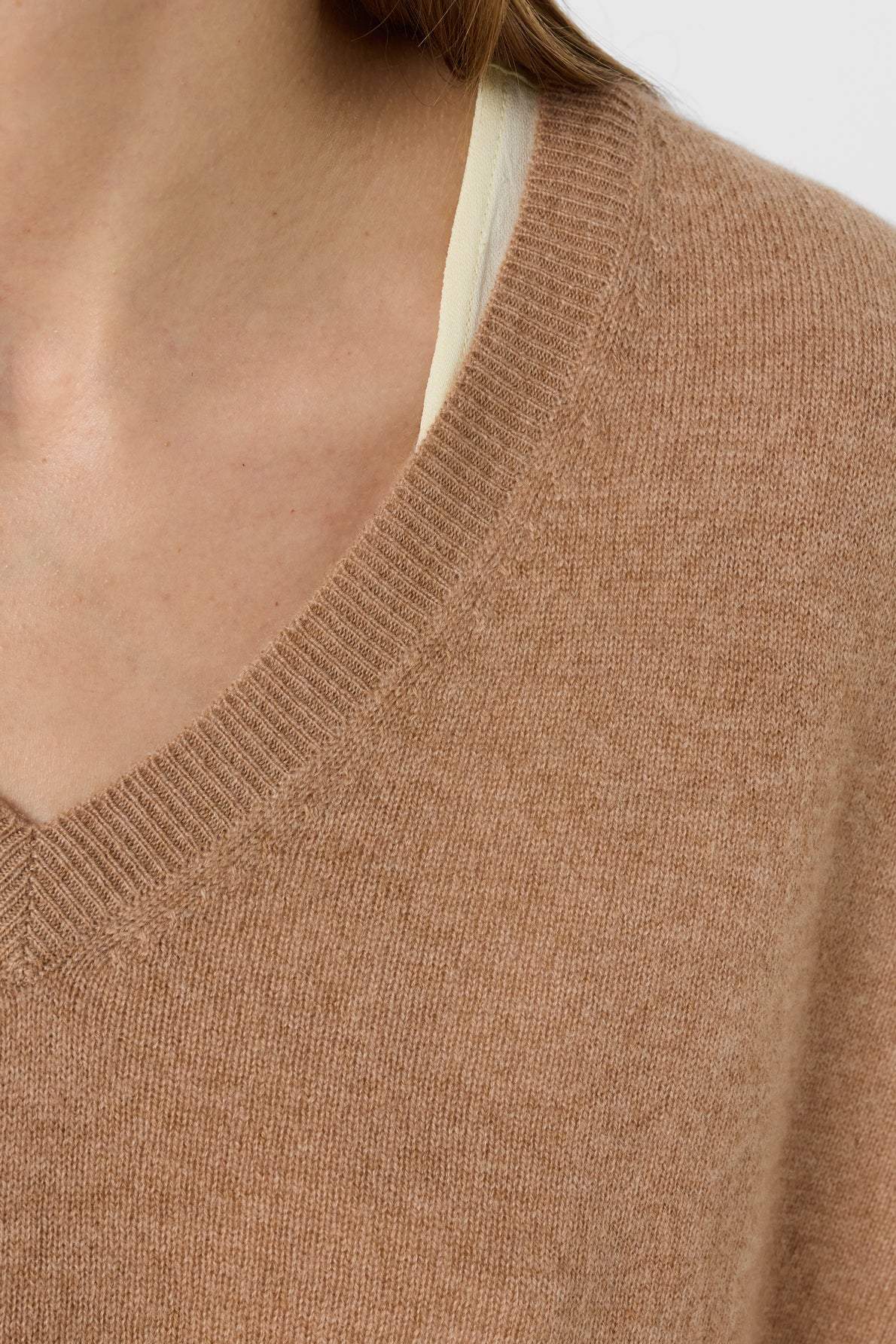 Close-up of a person wearing a light brown V-neck sweater with a cream-colored shirt or tank top underneath. Only the lower face, neck, and upper chest are partially visible.