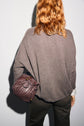 A person with wavy, reddish hair wears a taupe pull cachemire femme and holds a woven, brown handbag under their arm, facing away from the camera against a plain background.