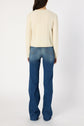 A person with long dark hair is standing facing away, wearing a pull écru en laine mélangée ornée de motifs géométriques and blue jeans with flared legs against a plain white background.