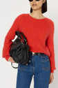 Woman wearing a bright red pull ample in laine mélangée, blue jeans, and sunglasses, holds a black leather handbag under her arm. The image is cropped above her knees and has a plain white background—a true touch of rouge baiser sophistication.