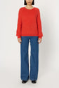 A person wearing a rouge baiser pull ample in laine mélangée, wide-leg blue jeans, and black shoes stands against a plain white background.
