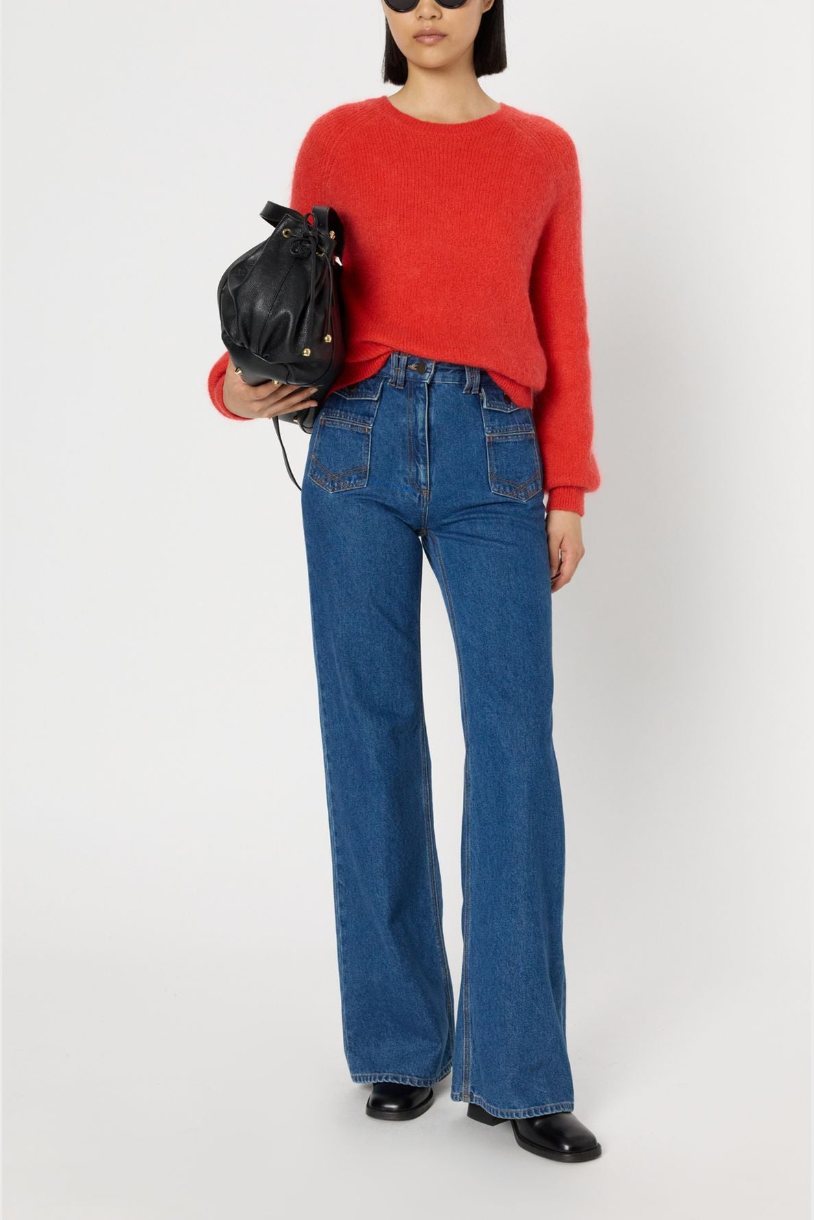A woman wearing a vibrant rouge baiser pull ample, high-waisted blue jeans, and black shoes stands holding a black handbag. She has straight dark hair and wears sunglasses. The background is plain white.