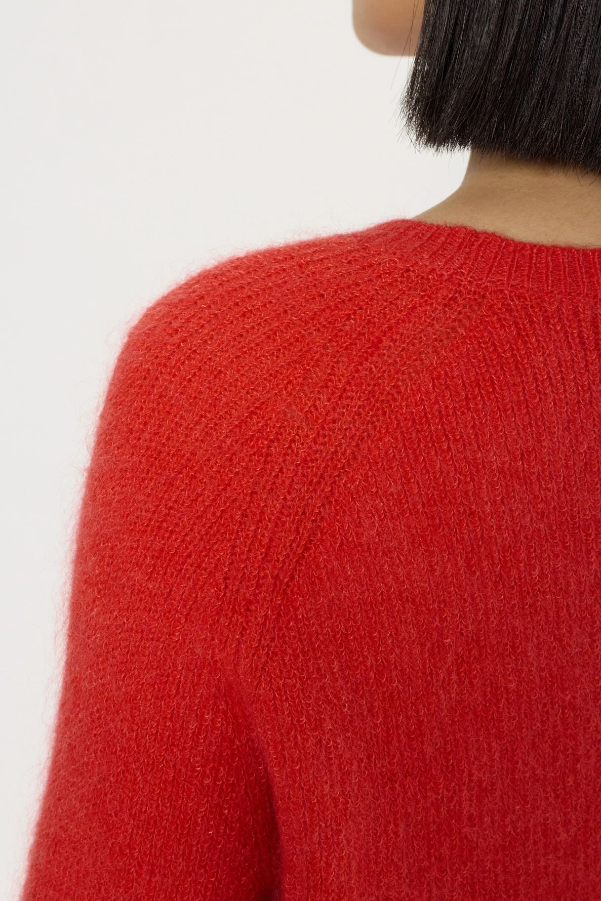 Close-up of a person wearing a fuzzy rouge baiser sweater in laine mélangée, showing the shoulder, upper back, and part of their head with short black hair. The background is plain and light-colored.