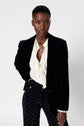 A person wearing a veste tailleur velours noire, a white ruffled blouse, and patterned black pants stands against a plain light background, looking confidently at the camera.