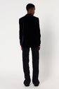 Person standing facing away from the camera, wearing a veste tailleur velours bleu marine and patterned trousers against a plain white background.