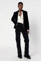 A person stands confidently wearing a veste tailleur velours bleu marine, a crisp white blouse, patterned dark pants, and black shoes against a plain white background.