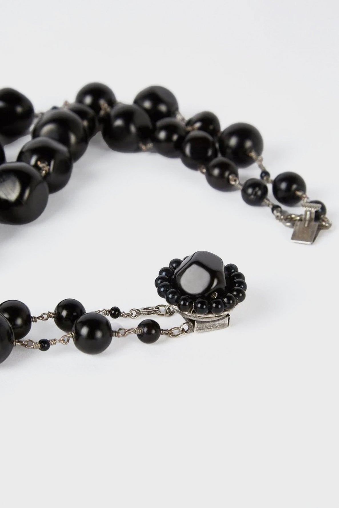 A close-up of a black beaded necklace with a decorative clasp featuring a central black stone surrounded by smaller black beads, laid out on a white surface.