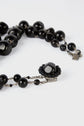 A close-up of a black beaded necklace with a decorative clasp featuring a central black stone surrounded by smaller black beads, laid out on a white surface.