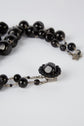A close-up of a black beaded necklace with a decorative clasp, featuring multiple round beads and a central glossy stone, laid on a light gray surface.
