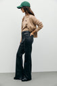 Person in a green baseball cap, tan cable-knit cardigan, beige top, and black pantalon taille haute flare stands in profile against a plain light background.