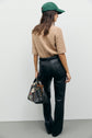 A person stands facing away, wearing a green cap, tan short-sleeve sweater, and a chic pantalon taille haute en cuir noir, holding a patterned handbag. Their hair is loose and wavy, and the background is plain white.