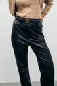 A person wearing a beige cable knit sweater and high-waisted black leather pants (pantalon cuir femme) with a black belt featuring a gold buckle, standing against a plain light background.