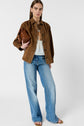 Woman standing against a plain background, wearing a brown jacket, light blue flared jeans, a white top, and sandals, holding a brown handbag under her arm.