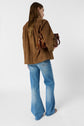 Person with long hair wearing a brown suede jacket and light blue flared jeans, standing and holding a brown leather handbag with a tassel, facing slightly away from the camera against a white background.